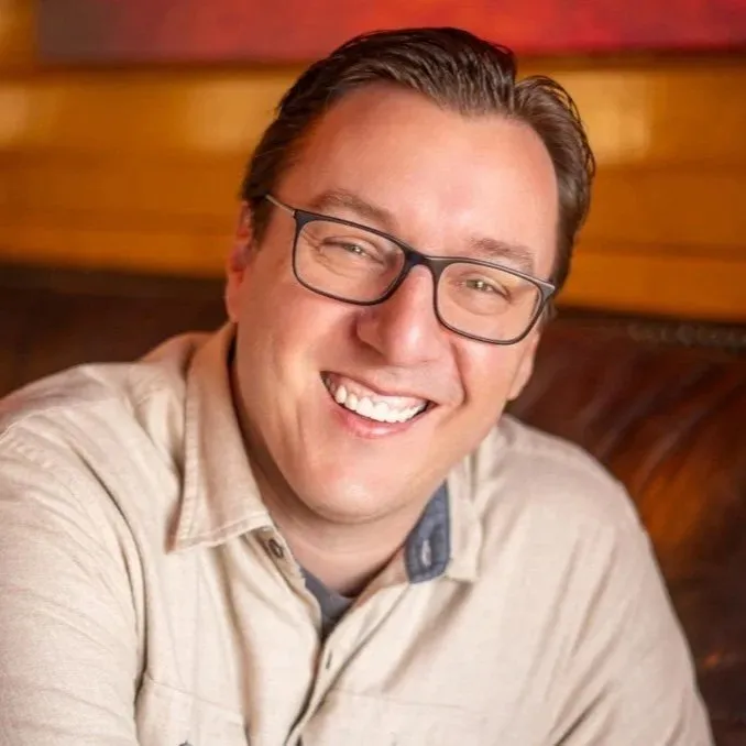 A smiling man with glasses and light brown hair, wearing a beige button-up shirt, sitting indoors against a wooden background.