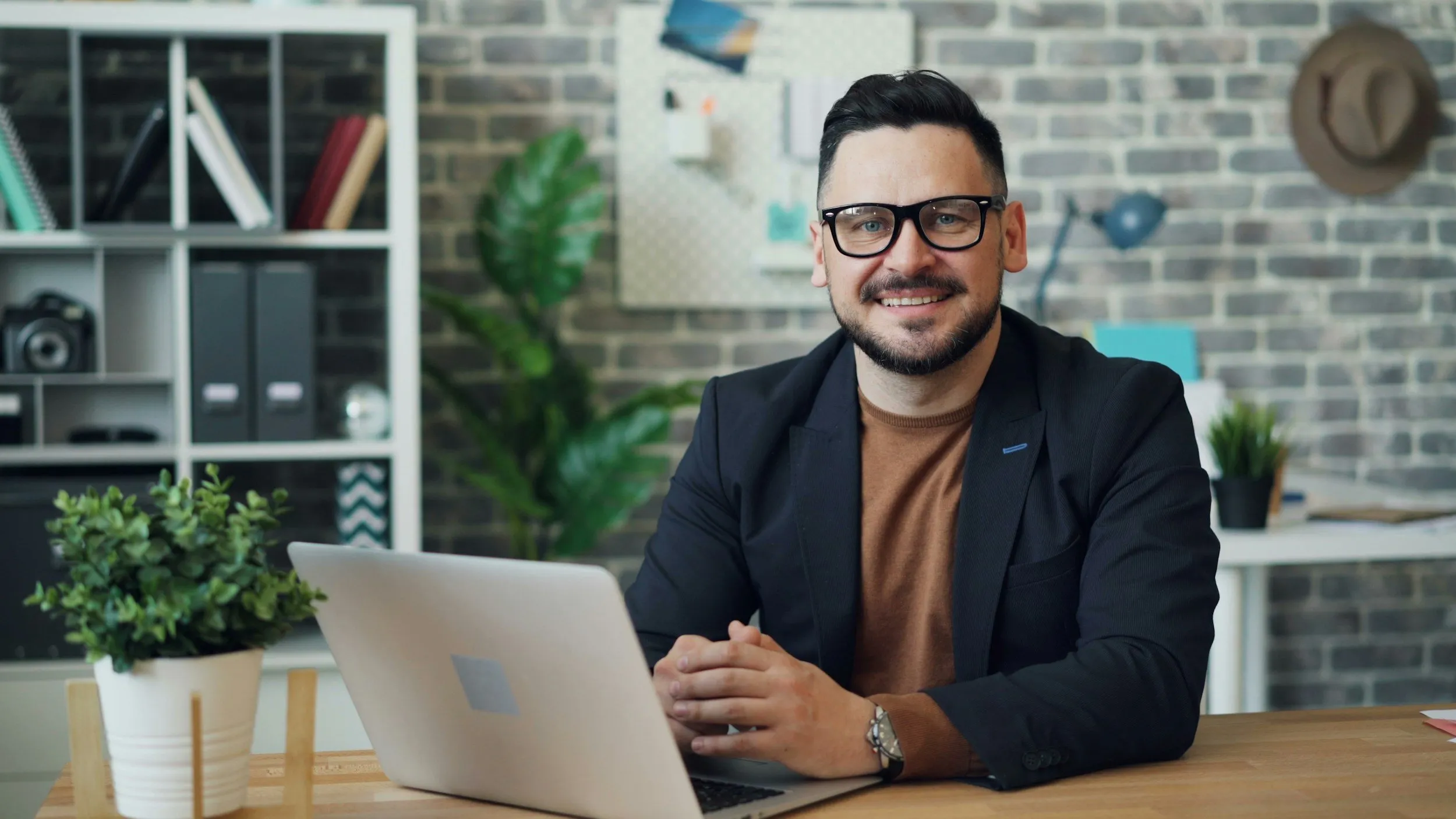 A man with glasses and a beard sitting at a desk in an office, smiling at the camera. There is a laptop in front of him, a potted plant on the desk, and a bookshelf with folders and books behind him.